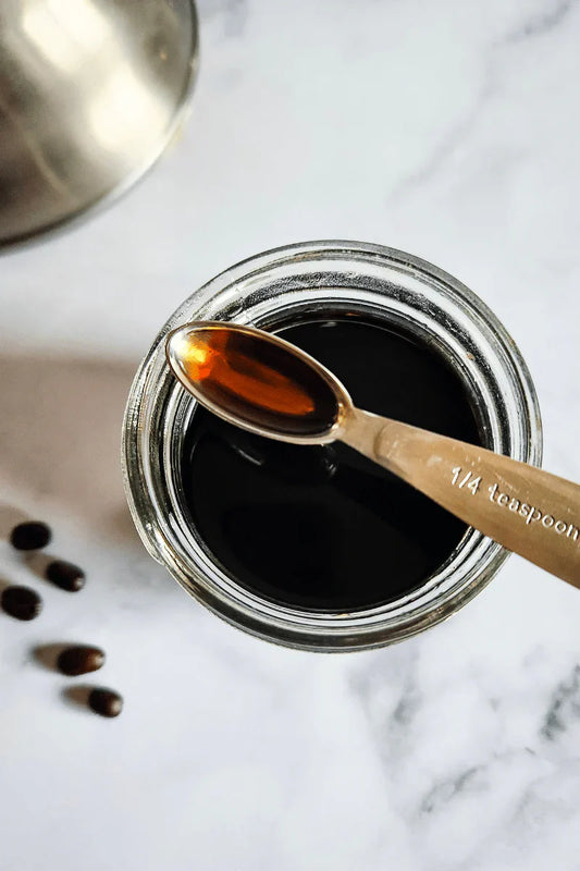 Frasco de cosmética natural con extracto de café sobre mesa de madera, junto a granos de café Magnagaea y hojas verdes.